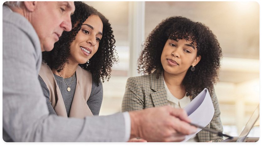 Grupo de profissionais em reunião, revisando documentos e conversando sobre informações apresentadas.